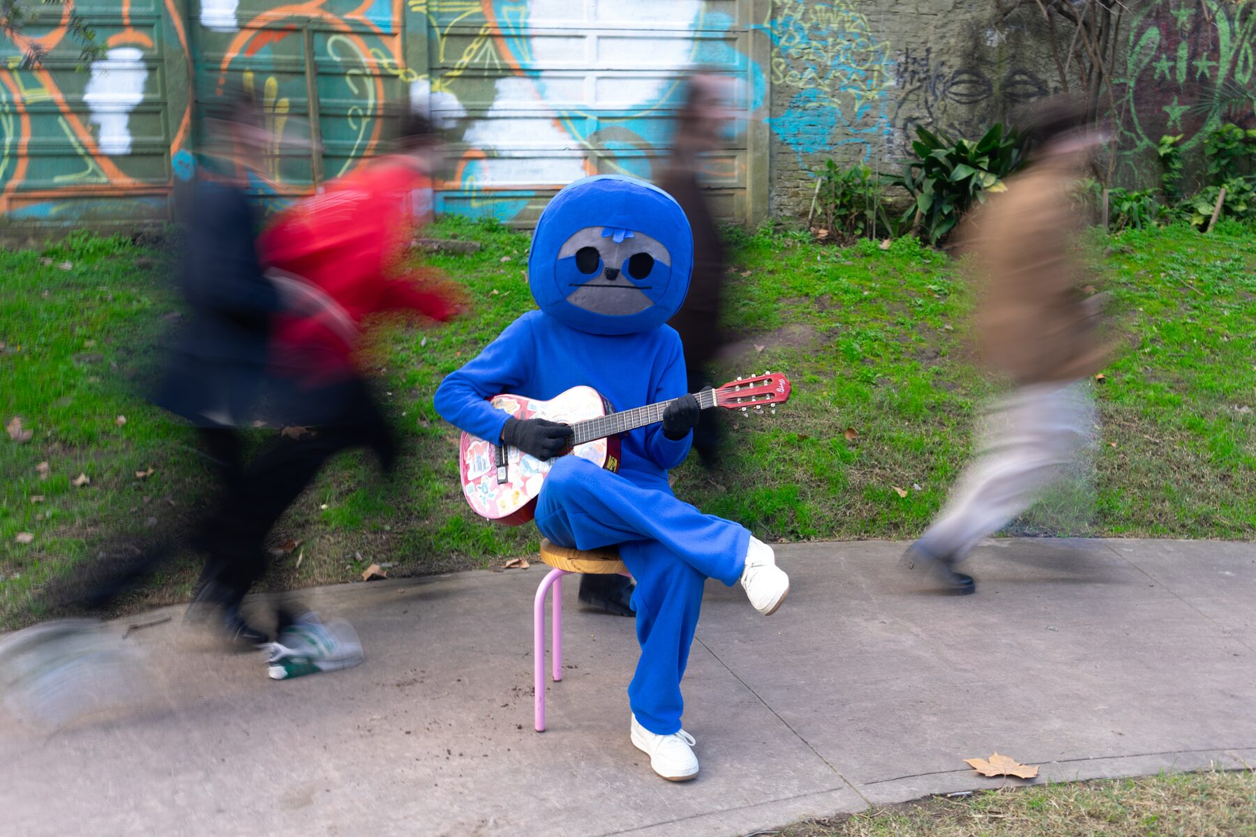 El perezoso celeste sentado en un banquito tocando la guitarra mientras cuatro personas pasan corriendo, borroneadas por el movimiento.
