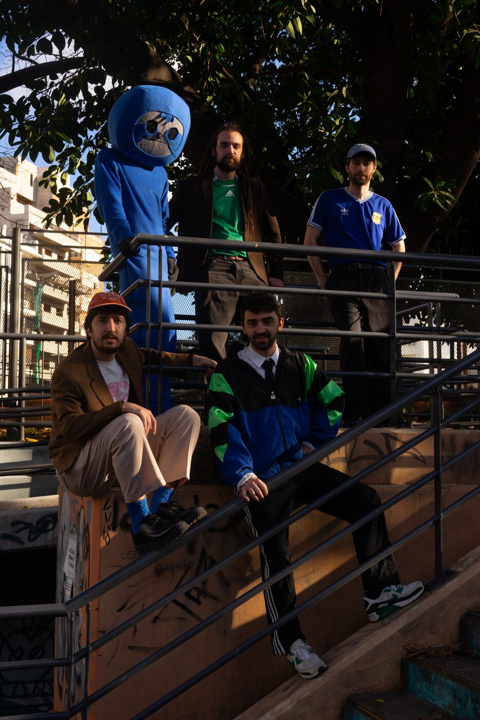 La banda y el perezoso en una escalera de barrio, entre rejas y árboles, con el sol de la tarde.
